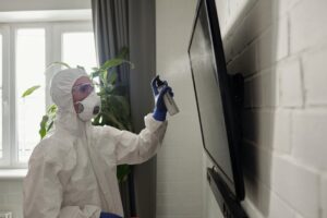 A person wearing full PPE disinfects a TV screen indoors, emphasizing health safety.
